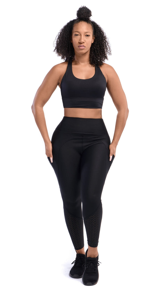 Front full view of a woman with a black sports bra and black leggings with her hands in both side pockets