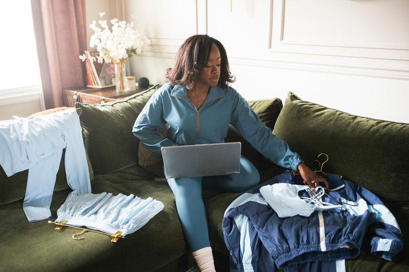Woman sitting on a couch with a laptop, surrounded by clothes, in a home setting.